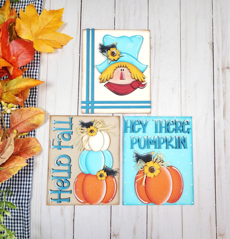 Three autumn-themed signs with a cartoon scarecrow, pumpkins, and text on a wooden surface.
