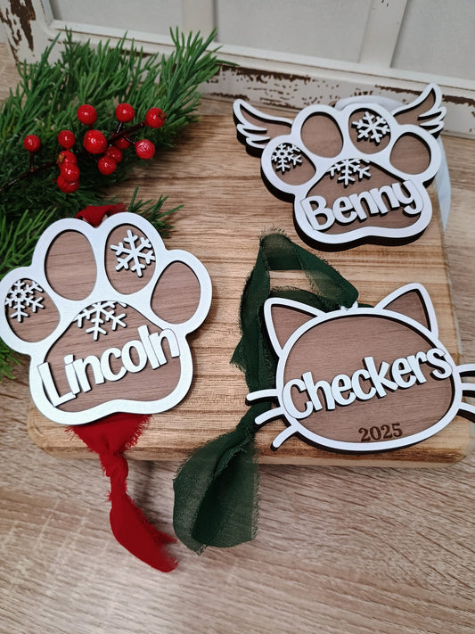 Three wooden ornaments shaped like paw prints and cat ears with names engraved on a wooden surface.